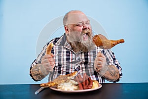 Hungry man with overweight eats chicken leg at table with fat food on blue background