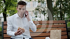 Hungry man in suit eating a hamburger when talking on phone with smile on bench