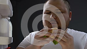 Hungry obese man chewing burger at night near fridge, overeating problem, diet