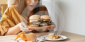 Hungry overweight woman holding hamburger on wooden plate, Fried chicken and Pizza on table .Concept of binge eating disorder BED photo