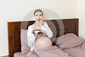 Hungry pregnant woman is eating a piece of tasty cake relaxing in bed at home. Sweet cravings during pregnancy