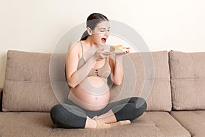 Hungry pregnant woman is eating a piece of tasty cake relaxing on the sofa at home. Sweet cravings during pregnancy