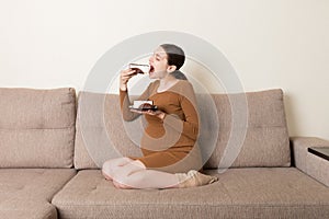 Hungry pregnant woman is eating a piece of tasty cake relaxing on the sofa at home. Sweet cravings during pregnancy