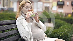 Hungry pregnant woman enjoying delicious burger sitting on bench in park