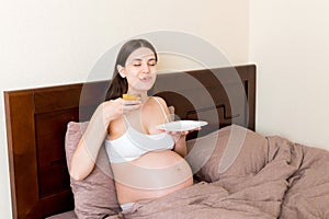 Hungry pregnant woman relaxing in bed is eating greedily a piece of cake and has a dirty mouth. Expecting mother can`t stop eatin