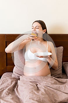 Hungry pregnant woman relaxing in bed is eating greedily a piece of cake and has a dirty mouth. Expecting mother can`t stop eatin
