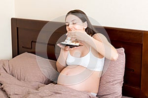 Hungry pregnant woman relaxing in bed is eating greedily a piece of cake and has a dirty mouth. Expecting mother can`t stop eatin