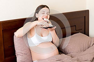 Hungry pregnant woman relaxing in bed is eating greedily a piece of cake and has a dirty mouth. Expecting mother can`t stop eatin