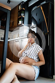 Hungry pregnant woman sitting on floor in kitchen longing for food
