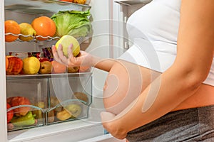 A hungry pregnant woman standing near refrigerator looking for healthy food during pregnancy.
