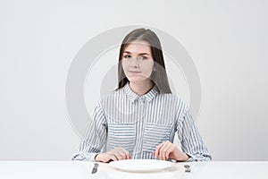 Hungry thin girl sitting at the table in front of an empty plate with a knife and fork.