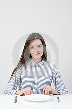 Hungry thin girl sitting at the table in front of an empty plate with a knife and fork.
