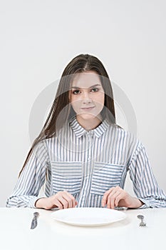 Hungry thin girl sitting at the table in front of an empty plate with a knife and fork.