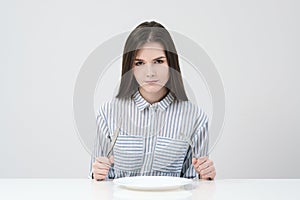 Hungry thin girl sitting at the table in front of an empty plate with a knife and fork.