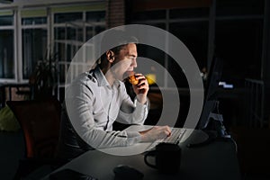 Hungry tired freelancer male eating burger working on desktop computer sitting on desk at office workplace late night in