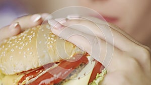 A hungry woman is eating burger at home kitchen. Big calories and cholesterol. Concept of fast food or unhealthy