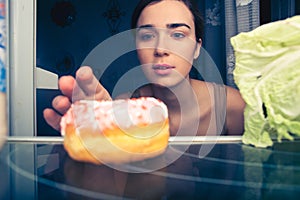 Hungry woman reaches for donut at night near fridge