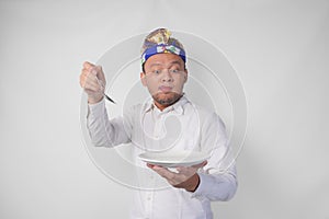 Hungry young Balinese man in white shirt and traditional headdress holding an empty plate with copy space and spoon while making a