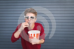 Hungry young man in 3D glasses eats popcorn from a cup and looks at the camera