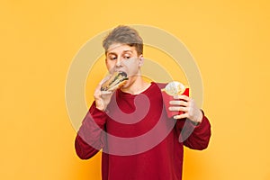 Hungry young man in casual clothing holds french fries in his hands and bites a burger on a yellow background. Guy teenager eats