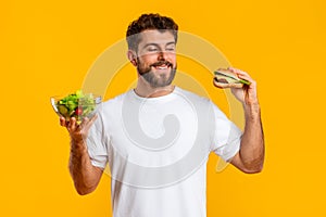 Hungry Young Man Choosing Between Burger And Salad, Yellow Backdrop