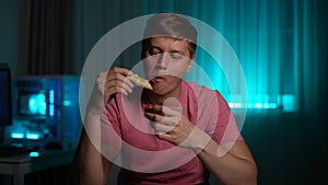 Hungry young man dipping slice of pizza in sauce sitting at table in evening or night on blurred background of soft