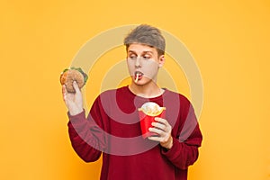 Hungry young man with french fries in his mouth, holding a burger in his hands and looking at him, isolated on a yellow background