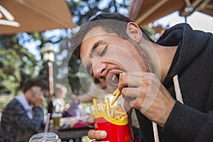 Hungry young man is greedily eating his french fries. He is sitting at the restauraunt terrace