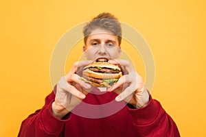 Hungry young man holds in the hands of a delicious big burger, looking at the camera on a yellow background. Focus on the sandwich