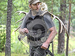 A hunter with a bow in the woods carries moose horns on his back and looking by side