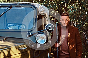 Hunter in elegant clothes holds a shotgun and standing a retro military car in the forest.