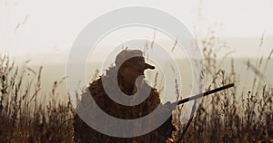 Hunter in hunting equipment lies in wait in the field, aims the target with shot gun foggy landscape on background