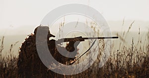 Hunter in hunting equipment lies in wait in the field. Silhouette of man in sunset light find the target and aim with rifle