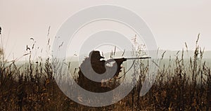 Hunter in hunting equipment lies in wait. Silhouette of man in sunset light find the target and aim with rifle foggy landscape