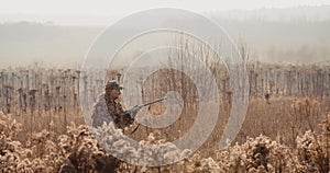 Hunter in hunting equipment with a rifle in his hand sneaks through the bush in the field, find the target and aim. Sunrise light