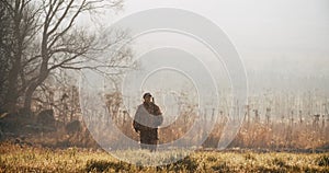 Hunter in hunting equipment with rifle on his shoulder walks through on camera in the field at foggy morning or sunny autumn