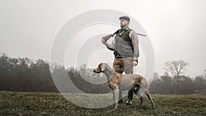 Hunter man with dog in traditional shooting clothes on field holding shotgun.