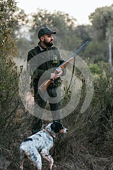 Hunter man with a gun standing outdoors with his dog while searching for a prey.