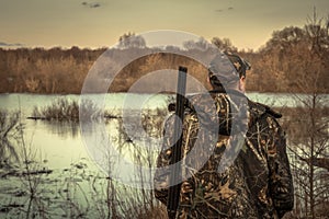 Hunter man shotgun camouflage exploring flood river hunting season rear view sunset