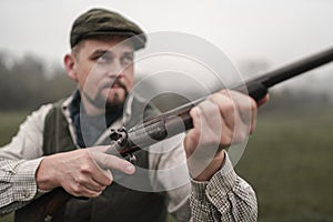 Hunter man in traditional shooting clothes on field aiming with shotgun.