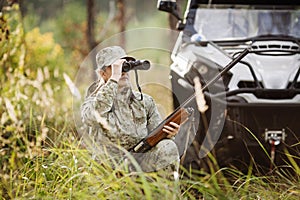 Hunter with shotgun looking through binoculars in forest
