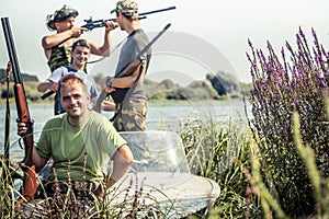 Hunters with guns in boat on river bank during hunting season among reed