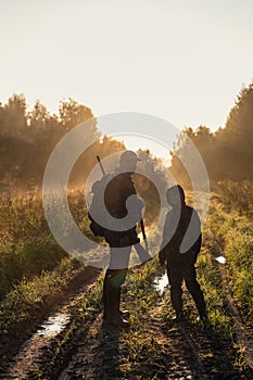 Hunters with hunting equipment going away through rural field towards forest at sunset during hunting season in