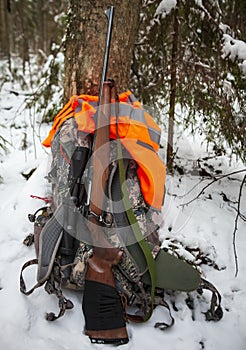 Hunting carbine stands at the trunk of tree next to backpack and red vest after roundup of winter hunting for ungulates