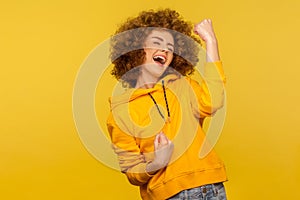 Hurray, I did it! Portrait of ecstatic enthusiastic curly-haired woman in urban style hoodie raising fists and screaming