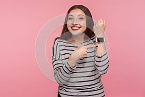 Hurry up! Portrait of positive woman in striped sweatshirt pointing smartwatch on her wrist and looking at camera