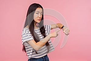 Hurry up, it`s late. Portrait of displeased angry girl with long brunette hair pointing at wrist watch