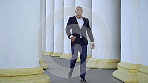 Hurrying African American man in suit walking between white columns outdoors hanging up smartphone and leaving. Wide