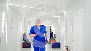 A hurrying older man doctor in a blue uniform runs along the bright corridor of the clinic holding a phone in his hands