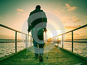 Hurt man with hooded jacket and forearm crutches standing on sea bridge